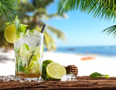 Glass of mojito with ice and lime garnishing below a palm tree in front of an Akumal beach.